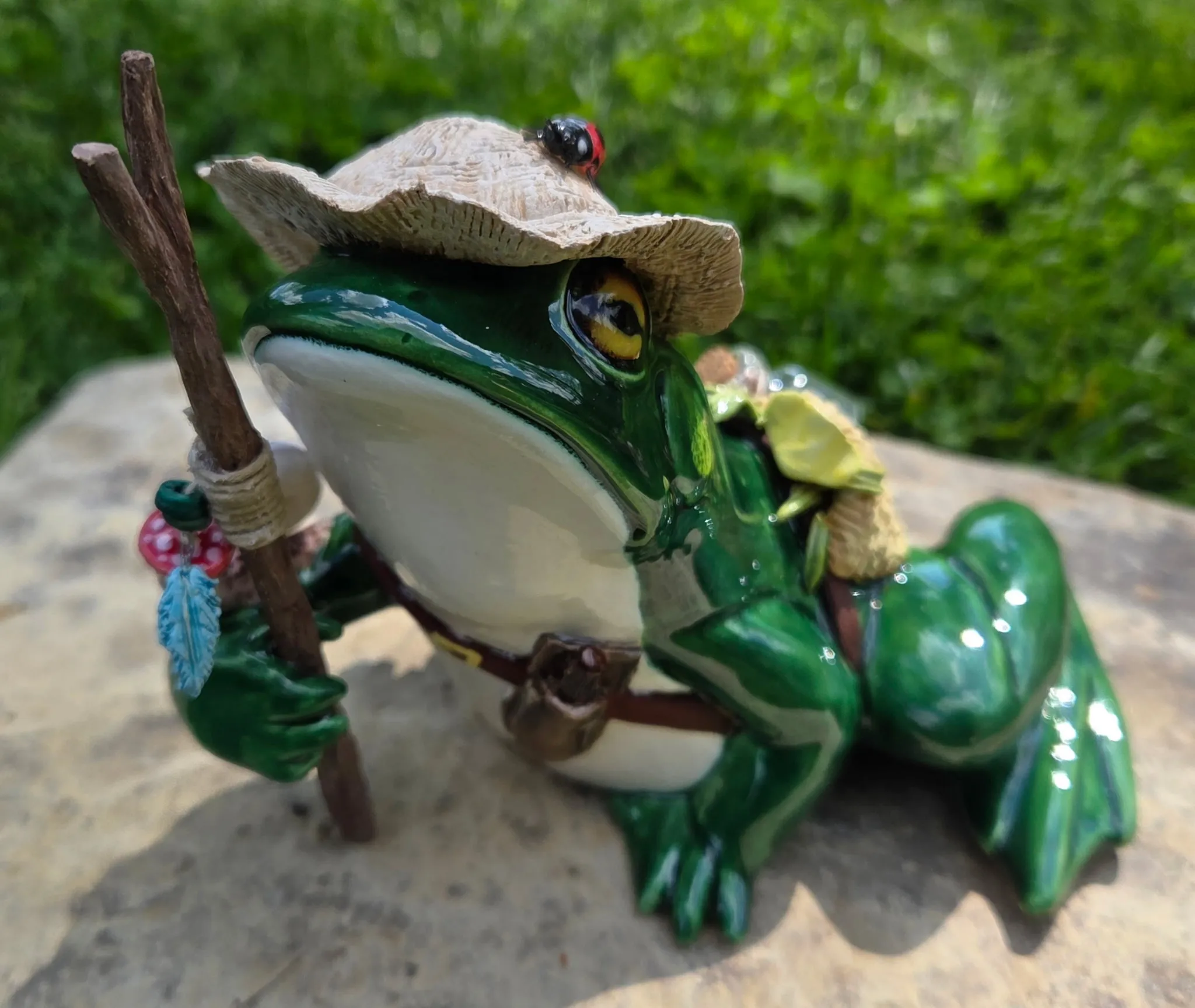 Green-glazed ceramic frog wearing a tiny straw hat with a ladybug on the brim, holding a wooden walking stick, with a small woven backpack, sitting on mossy stone.