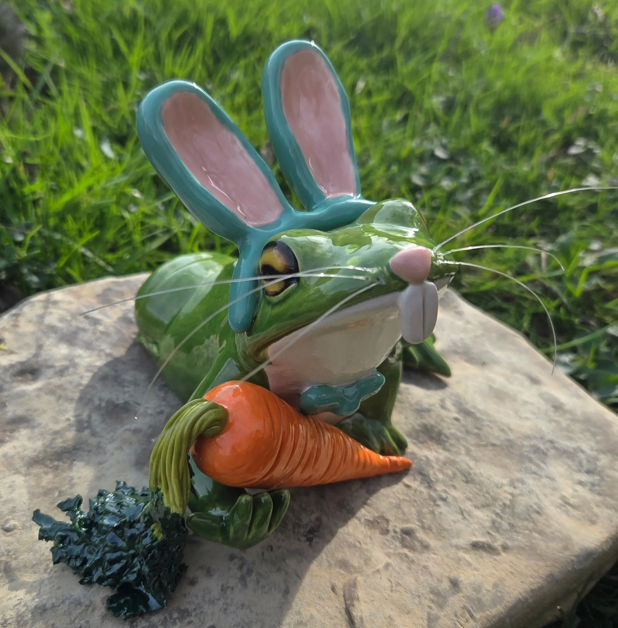 Green-glazed ceramic frog wearing teal bunny ears, holding a large orange ceramic carrot, sitting on a flat stone in grass.