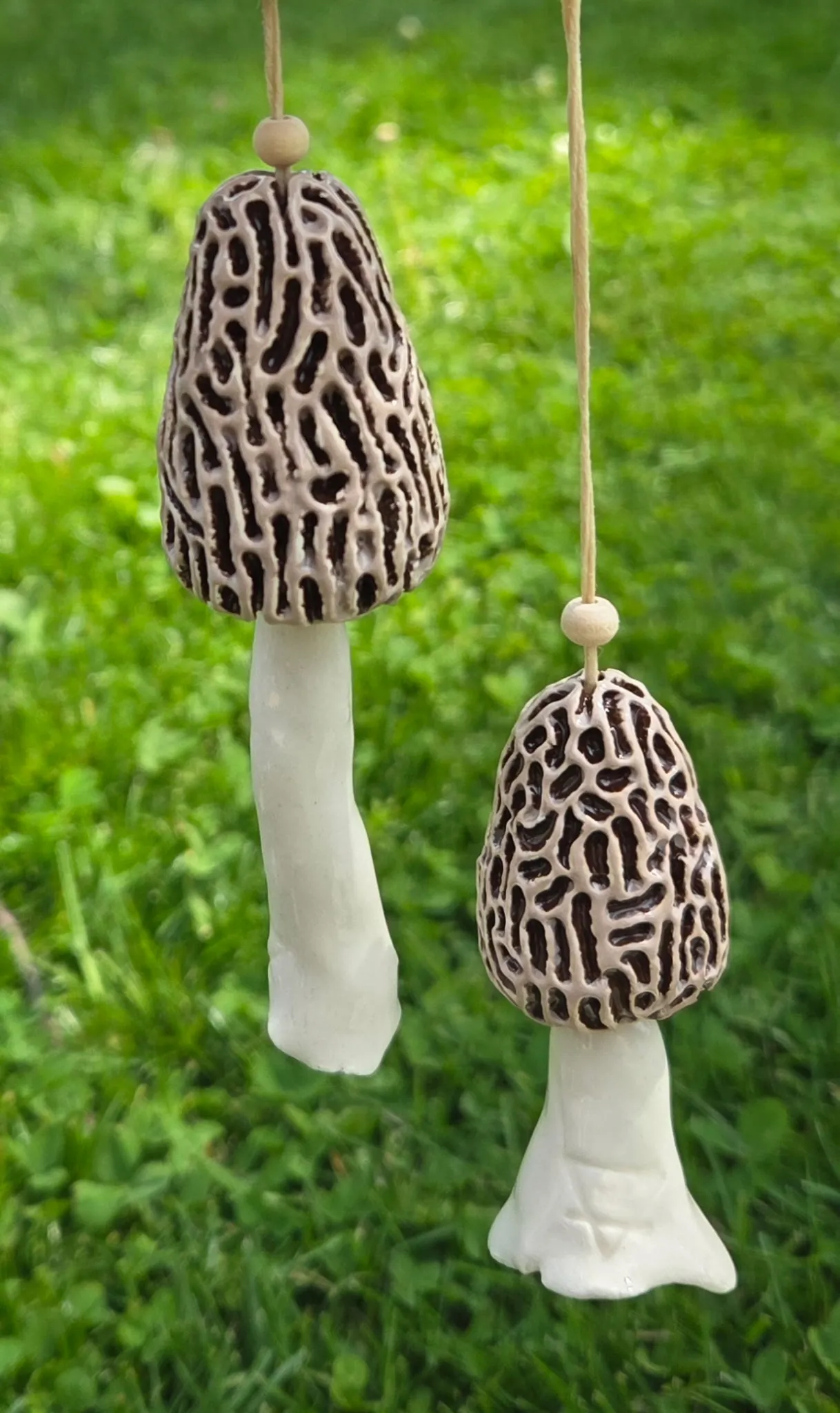 Pair of hanging ceramic morel mushrooms with honeycomb-textured brown caps on pale stems, strung on natural cord with wooden beads, against a blurred grass background.