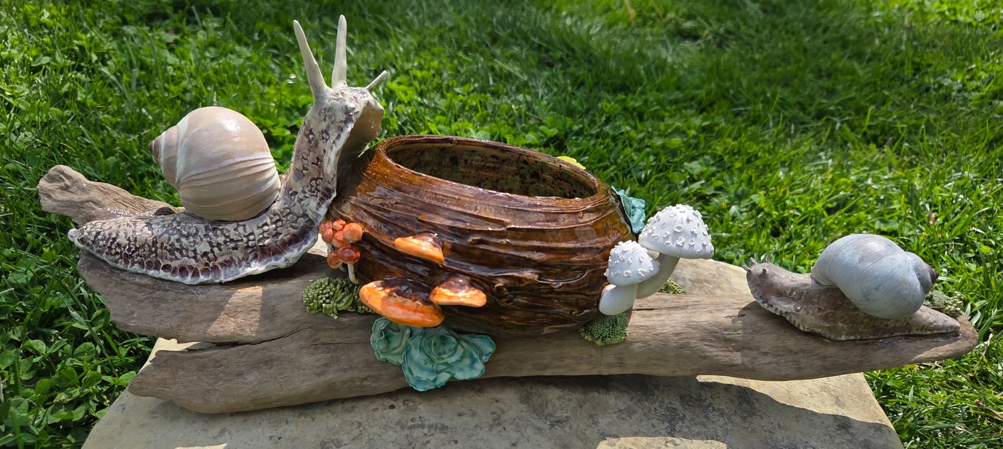 Ceramic garden assemblage on driftwood — a large speckled snail, a small hollow-log bowl with painted orange mushrooms clustered around it, and a smaller snail on the far end.