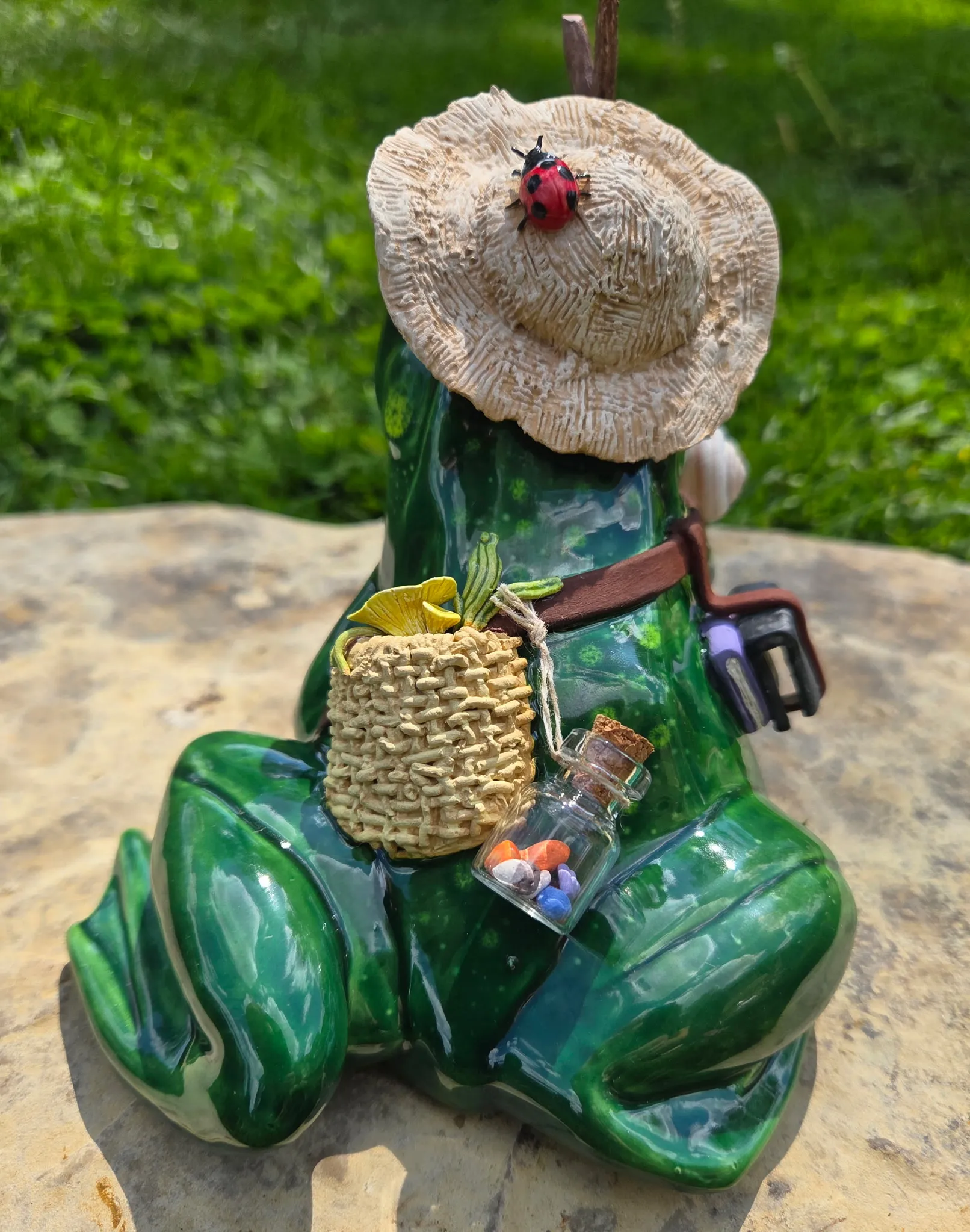 Back view of a green-glazed ceramic frog with straw hat, showing a woven-textured backpack holding a small ceramic specimen jar of painted beetles.
