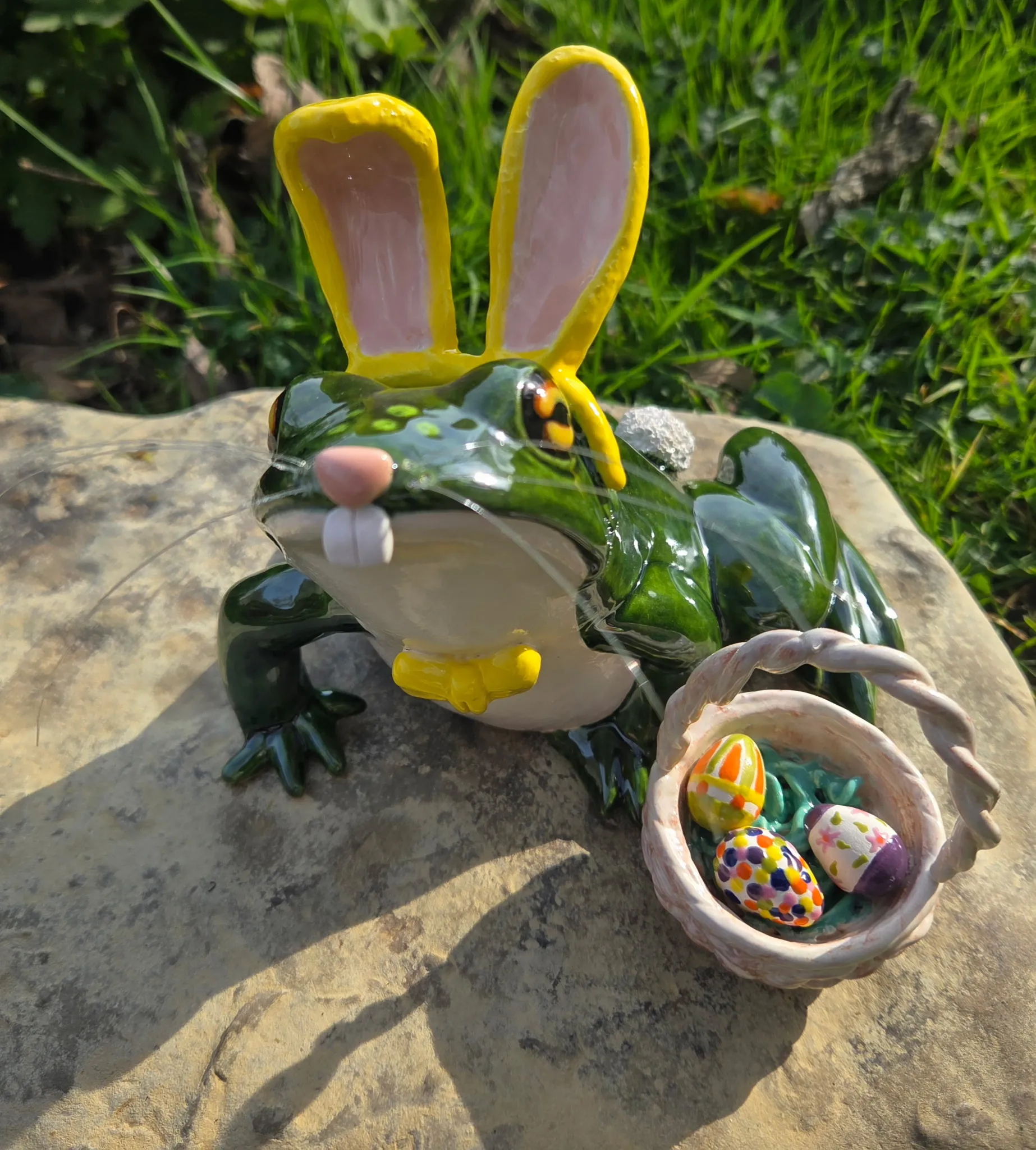 Green-and-yellow ceramic frog with yellow bunny ears, beside a small ceramic basket filled with painted Easter eggs, on stone in grass.