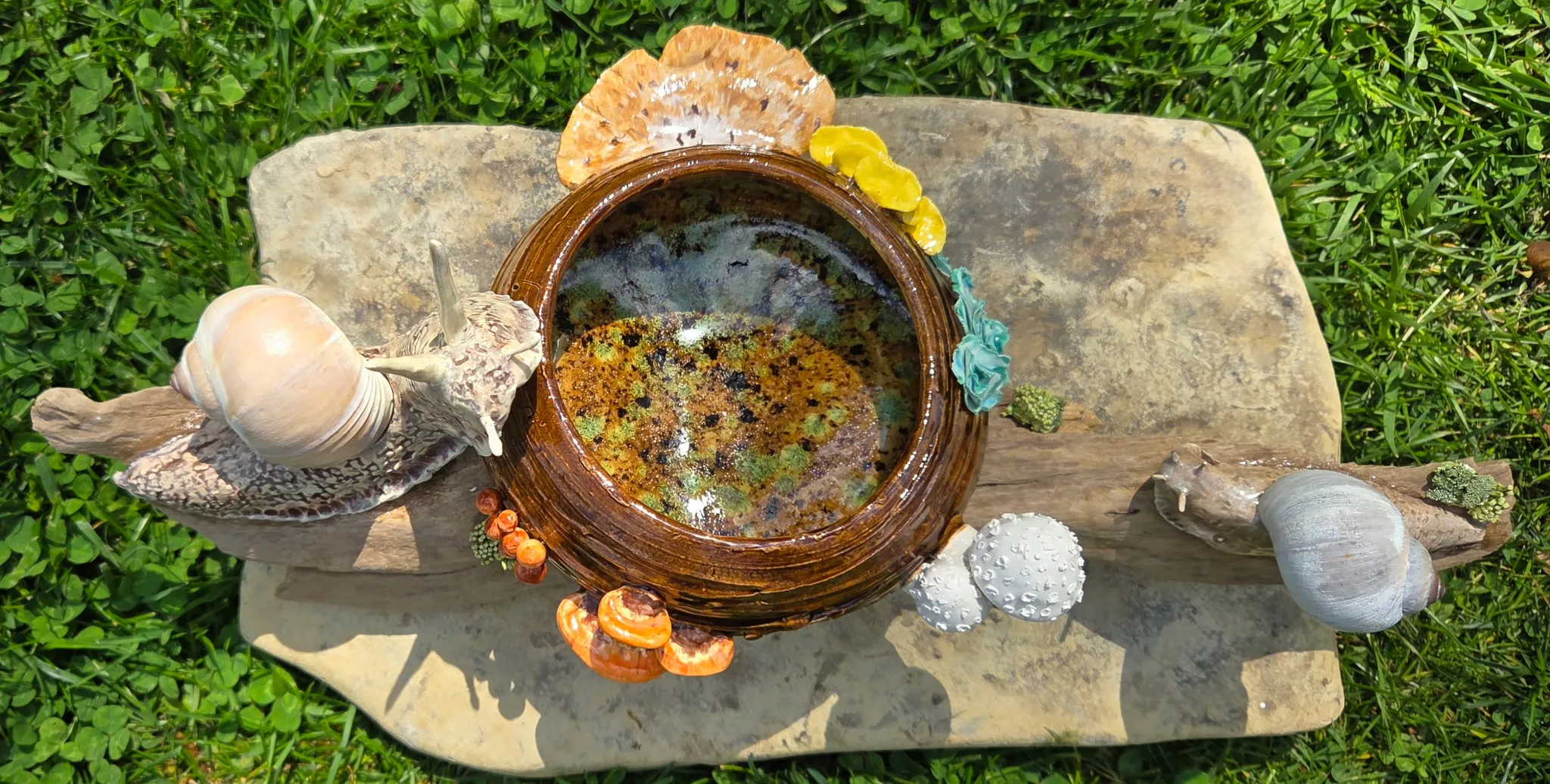 Driftwood piece with a hand-thrown glossy brown ceramic bowl at its center, surrounded by small ceramic snails, mushrooms, and a pale moon-like stone.