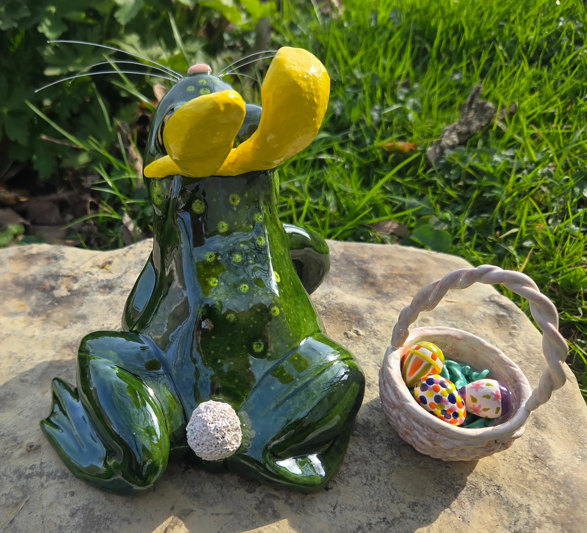 Green-glazed ceramic frog seen from behind, yellow sunflower on his back, accompanied by a small woven-textured ceramic basket of decorated eggs.