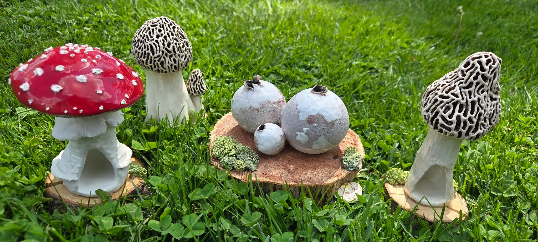 Set of ceramic mushrooms — a red-and-white-spotted fly agaric, two brown honeycomb morels, and a small cluster of speckled painted eggs — displayed on a wooden log slice in grass.